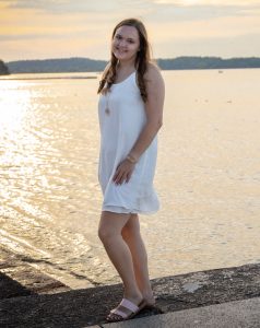 A women in a white dress standing on a pier in front of water and a sunset