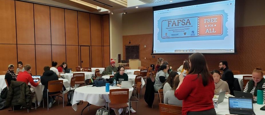A large room with people sitting around round tables, a few looking at laptop, or up at a screen that shows the visible text 'FAFSA Free for All"